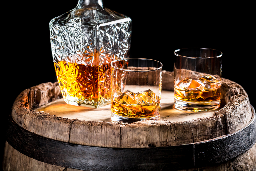 En bägare och två glas fyllda med whiskey ovanpå en rustik tunna